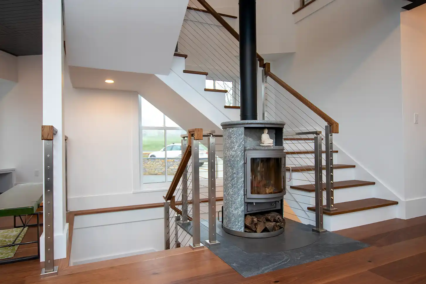 Cylindrical wood stove wrapped in stone beside a cable-rail staircase